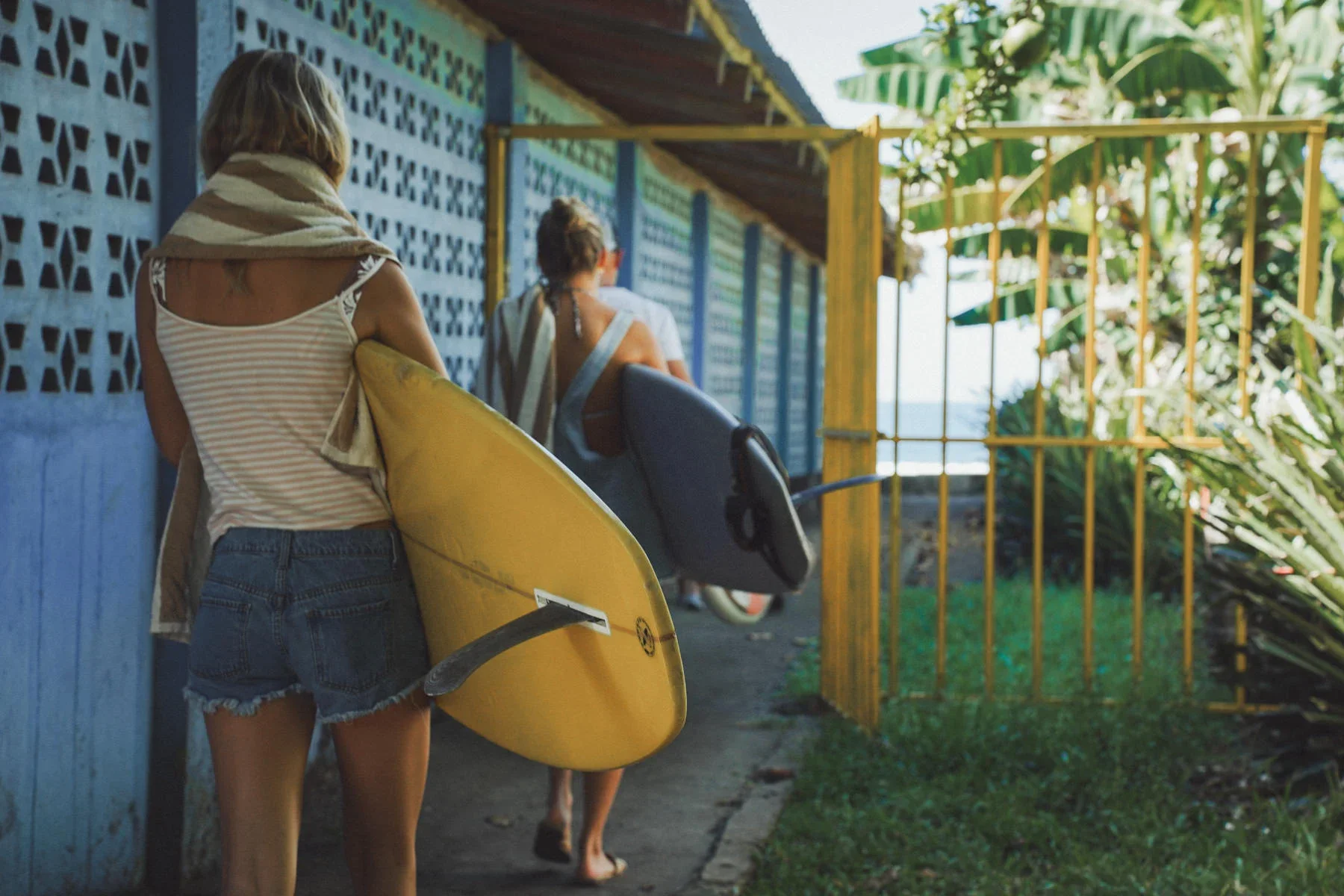 Two surfers heading to the beach with surfboards during a surf trip in Costa Rica, tropical plants and colorful walls in the background.