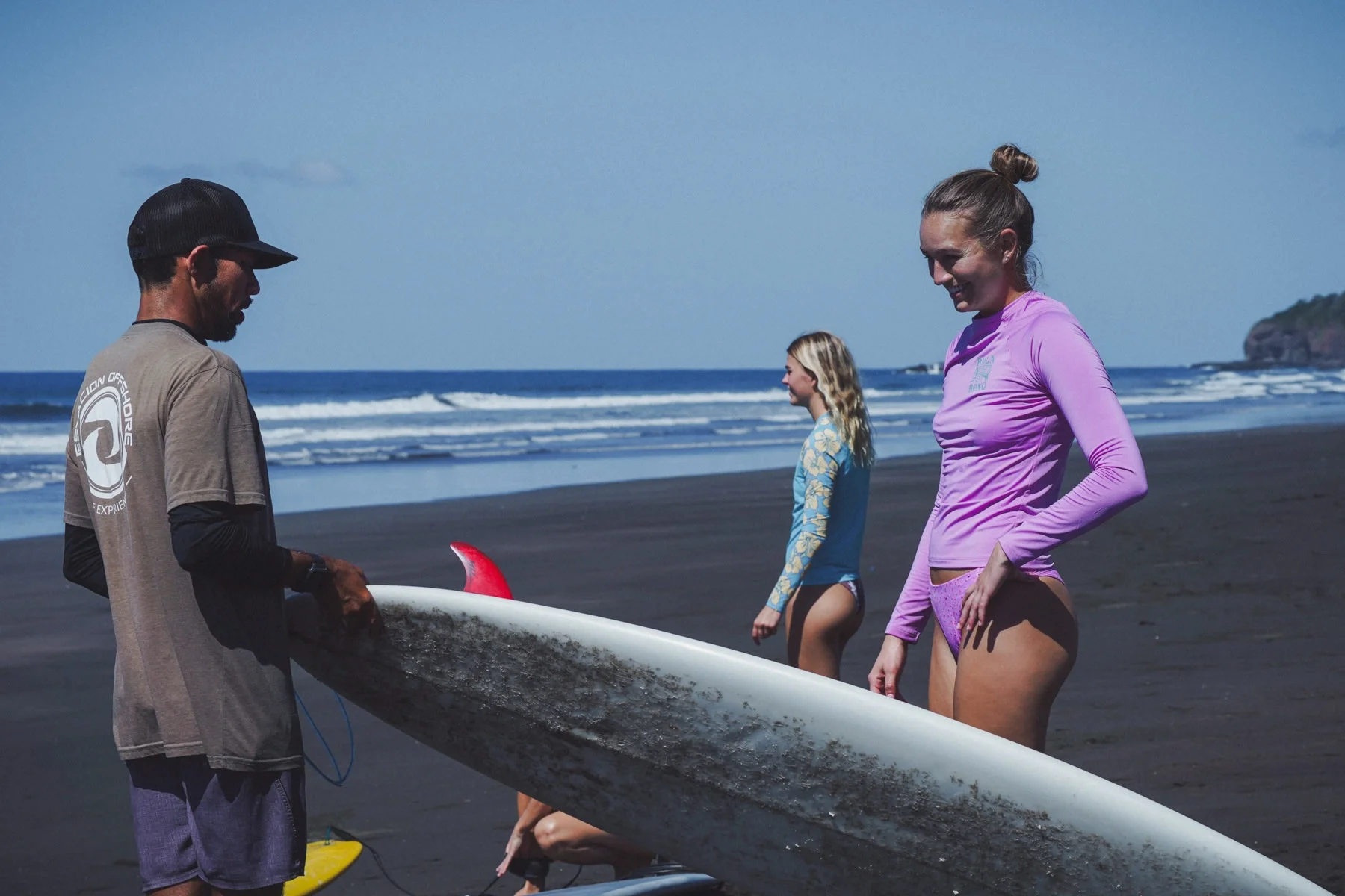 ISA-certified surf coach with two students on the black sand beach of Playa Ostional Costa Rica, ocean waves and rocky outcrop in the background