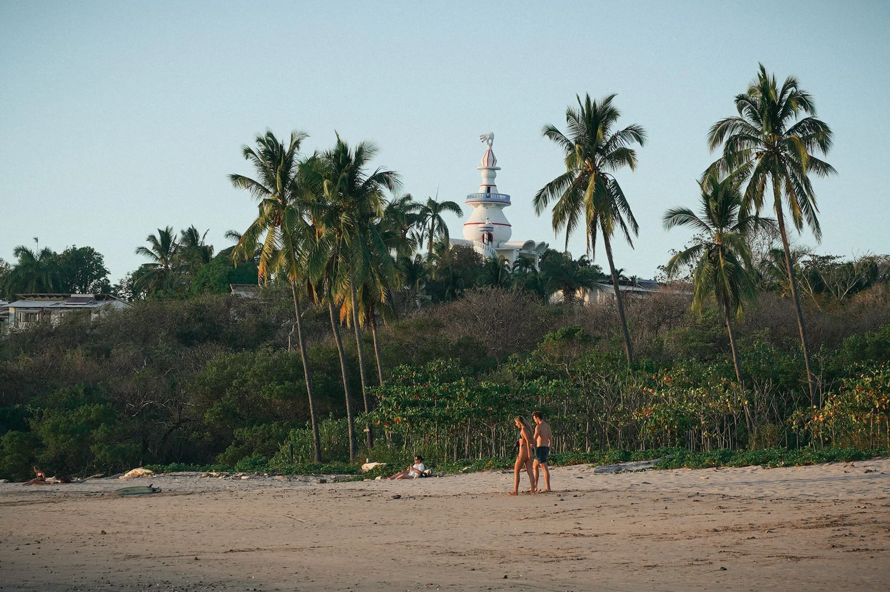 Playa Guiones beach scene with a couple walking on the sand, palm trees, and Nosara Beach Hotel in the background.