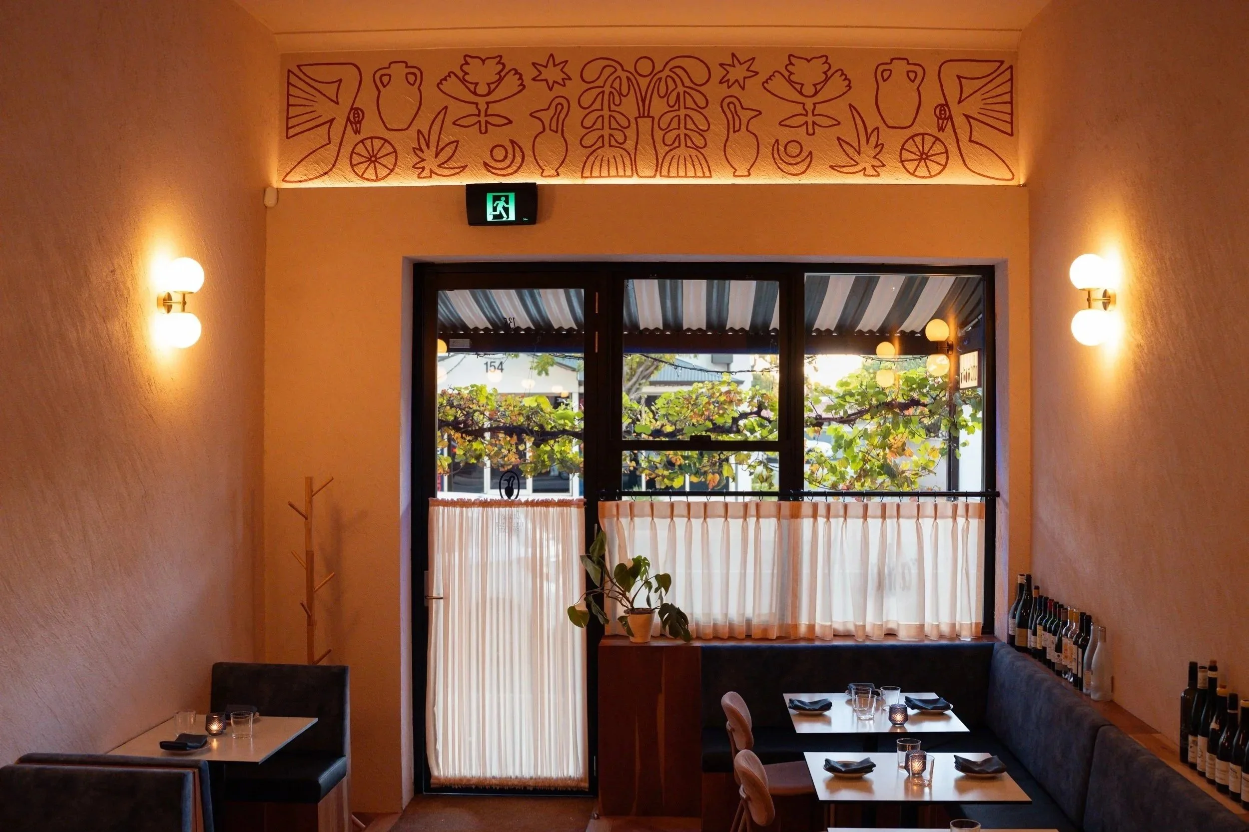 Interior of a cozy restaurant dining area with two tables set with glasses, black napkins, and small candles. The room has a window with outdoor greenery, soft lighting with wall sconces, and a decorated wall with abstract art above the window. Bottles are lined up on a shelf on the right side.