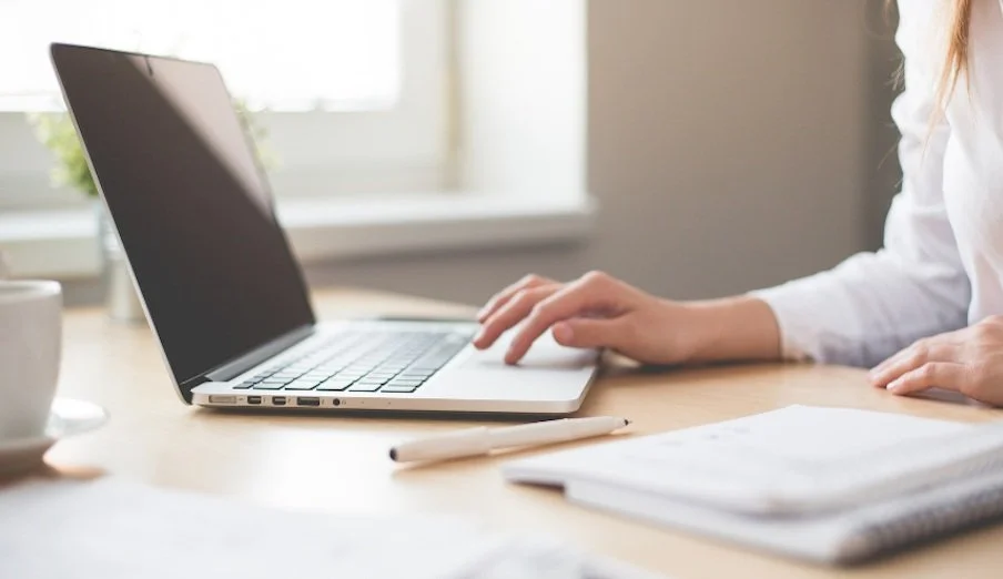 Person working on a laptop at a desk with a notebook and pen nearby