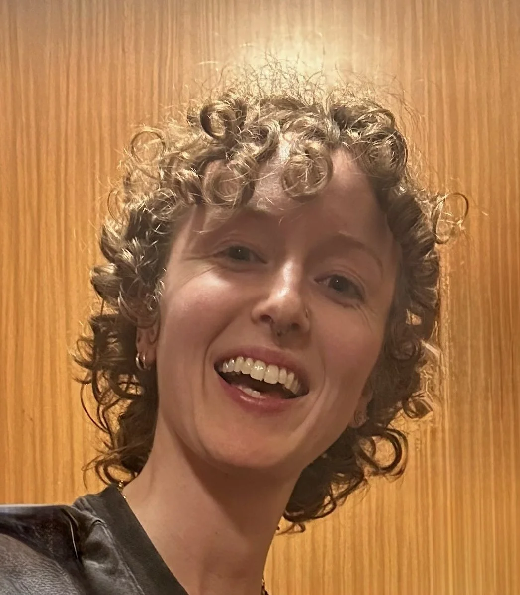 A woman with short, curly hair, smiling with her mouth open, standing in front of a wooden door