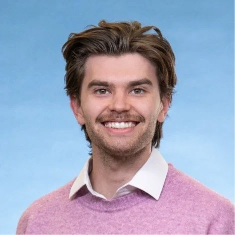 A man smiling with short brown hair, light facial hair, wearing a collared shirt underneath a sweater, standing in front of a blue background