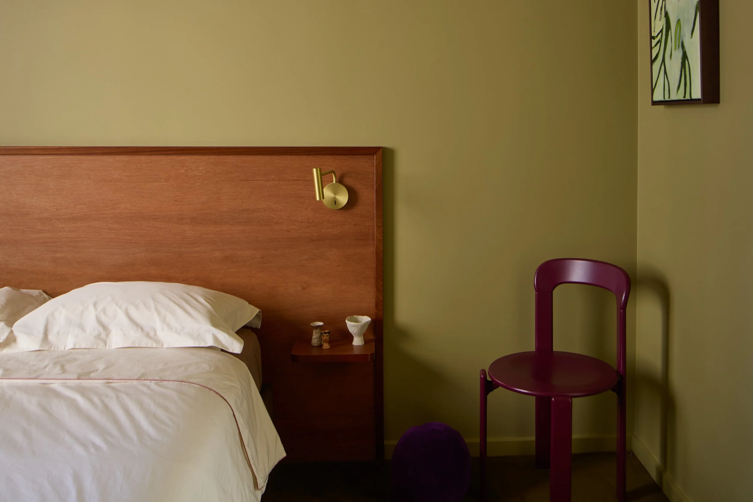 A bedroom corner with a wooden bed and white bedding, a small side table with decorative items, a purple chair, and wall art.