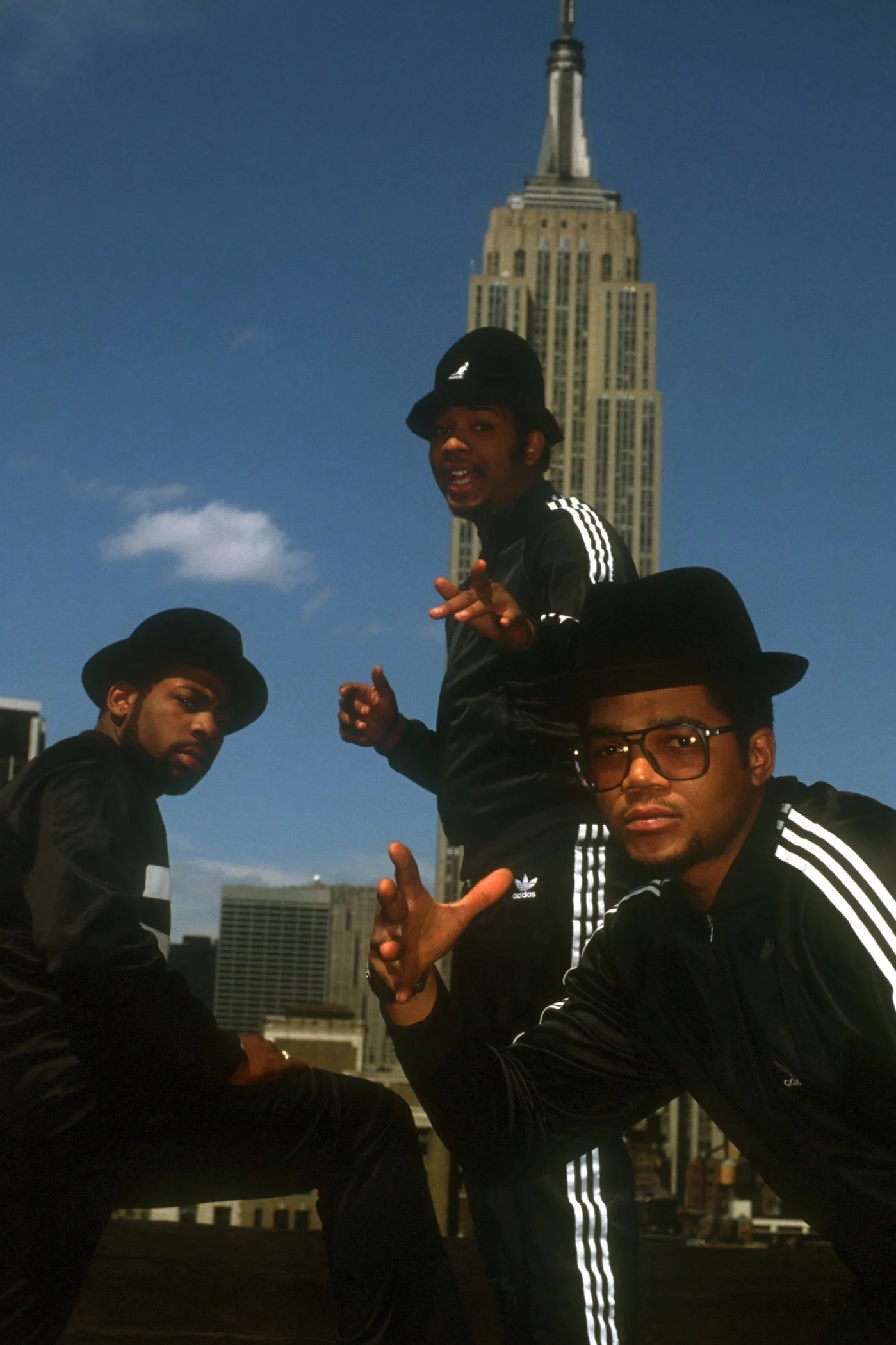 Three young men dressed in black Adidas tracksuits and black hats posing in front of the Empire State Building in New York City.