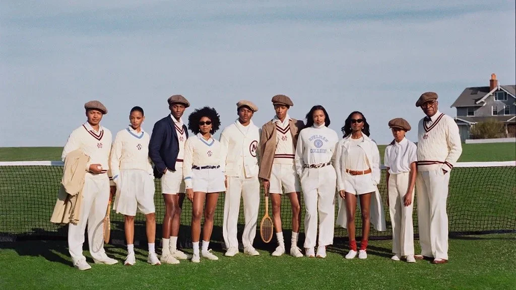 A group of ten young people dressed in vintage tennis attire, standing on a grass court with tennis rackets, in a suburban neighborhood setting.
