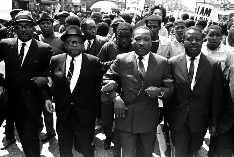 Civil rights leaders marching arm in arm during a protest in the 1960s, with a crowd behind them, some holding signs.