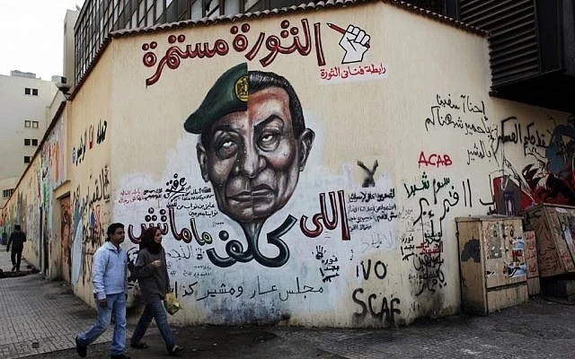Street mural depicting a figure in a military hat with Arabic writings, political graffiti on a building wall, and two people walking by, urban setting.