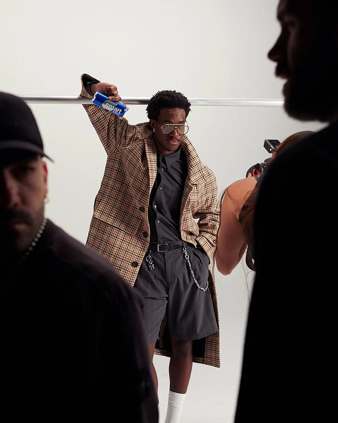 A male model with glasses and a plaid coat striking a pose in front of a white background, surrounded by photographers.
