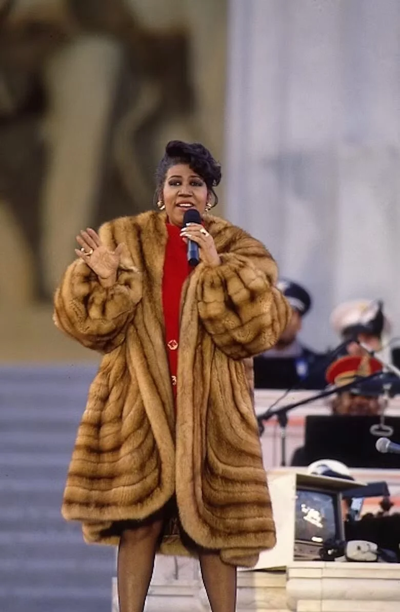 A person wearing a luxurious fur coat sings on stage, holding a microphone, with musicians in the background.