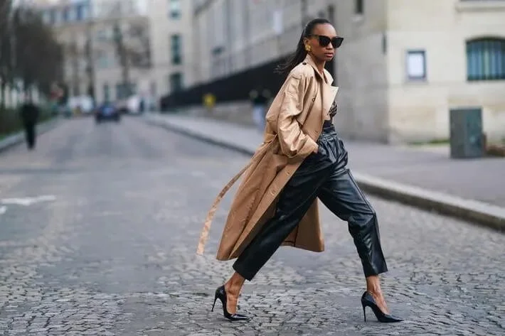 Fashionable woman wearing a tan trench coat, black leather pants, and high heels, walking confidently on a cobblestone street. She is also wearing sunglasses.