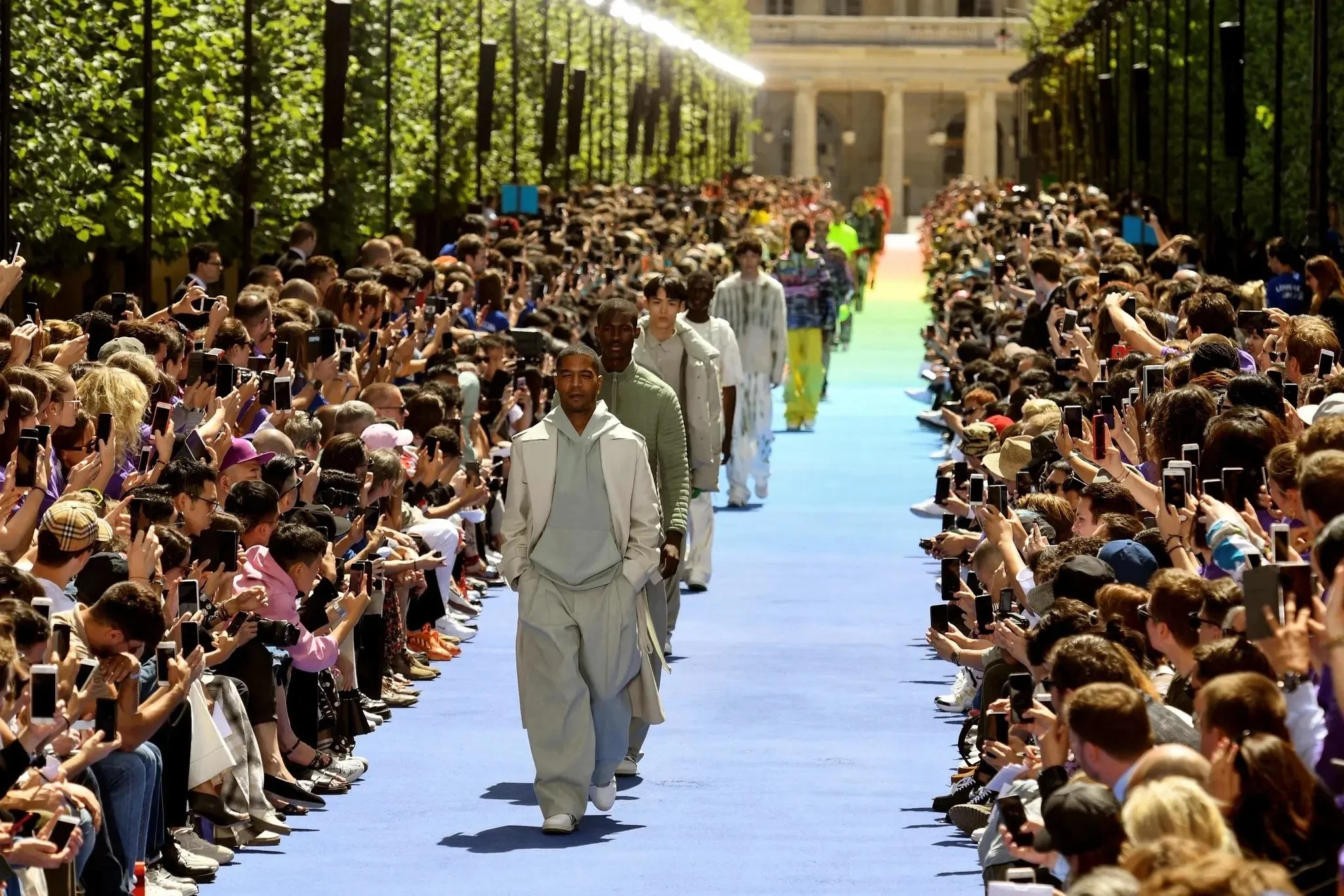 Models walk down a long runway at a fashion show, surrounded by a large audience taking photos, under a canopy of trees with sunlight filtering through.