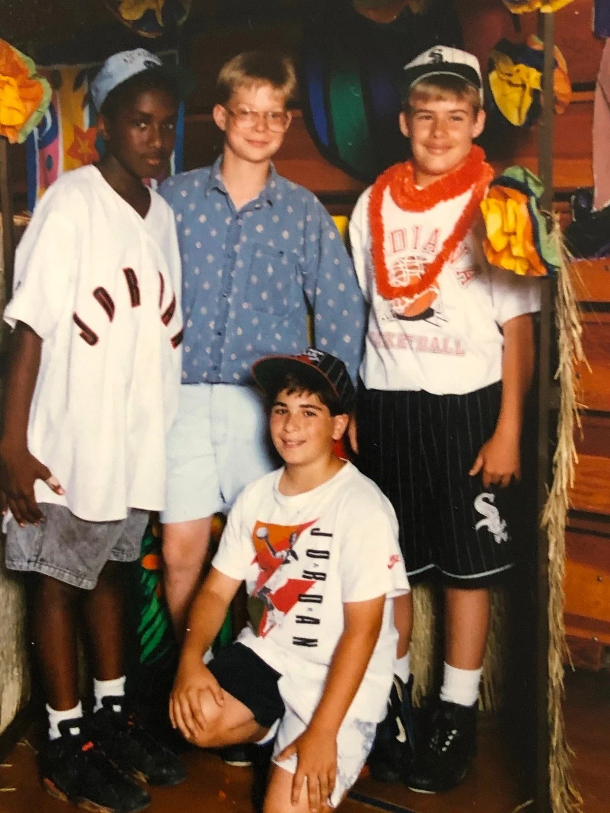 Four young boys posing together, one kneeling, wearing casual clothing including a "Jordan" t-shirt and sports apparel. The setting has colorful decorations and a wooden backdrop, suggesting a festive or themed event.