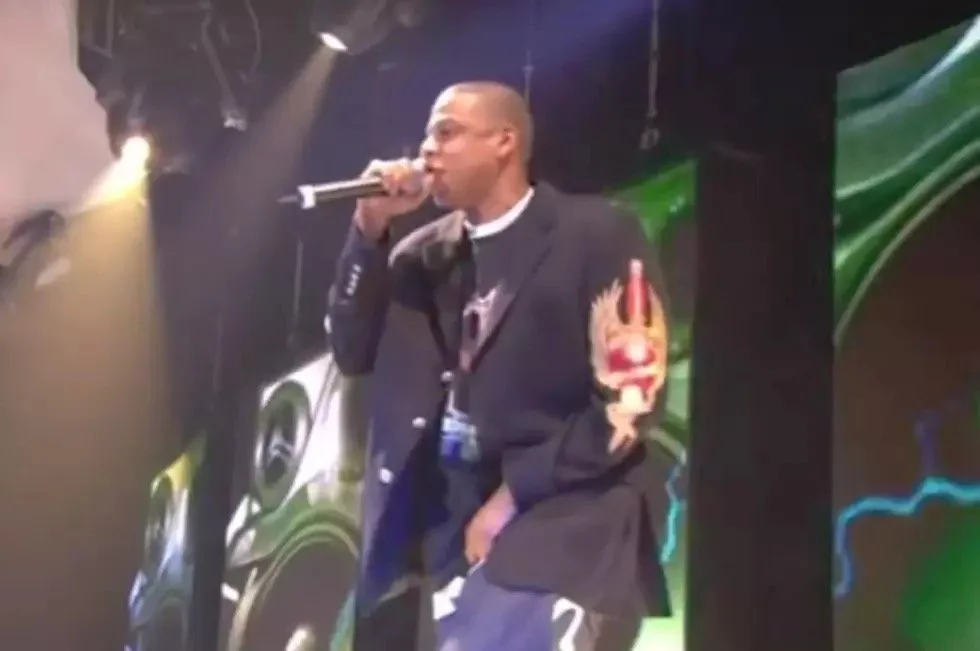 Man in a suit jacket with a patch on the sleeve, holding a microphone, performing on stage with digital visuals in the background.