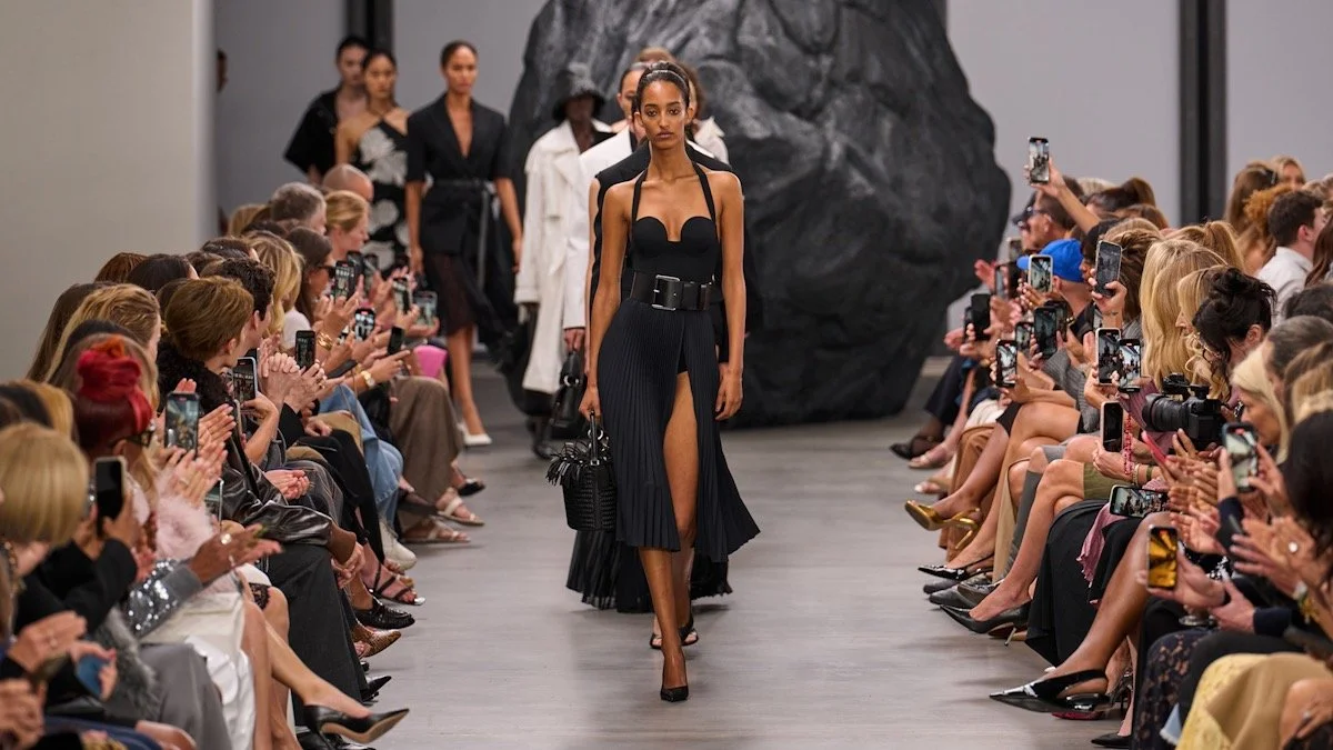 Fashion runway show with models walking down the runway, audience on both sides taking pictures, and a large black rock prop in the background.