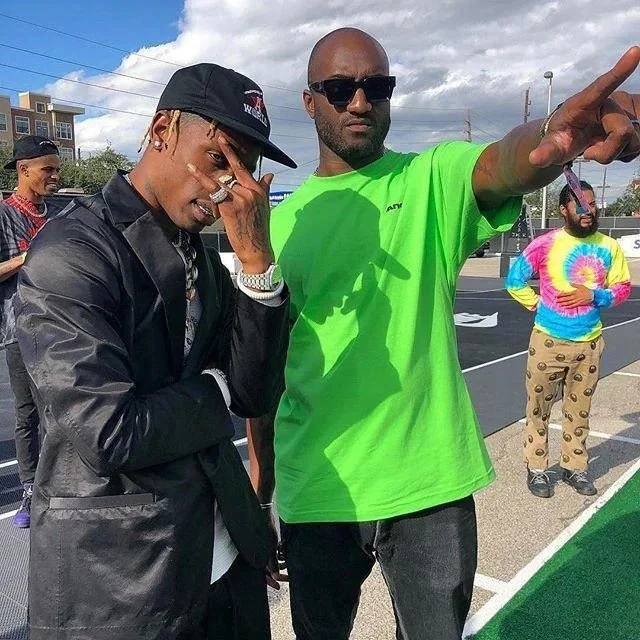 Group of people outdoors with one man pointing and another wearing a black hat and jacket, bright green shirt, and sunglasses.