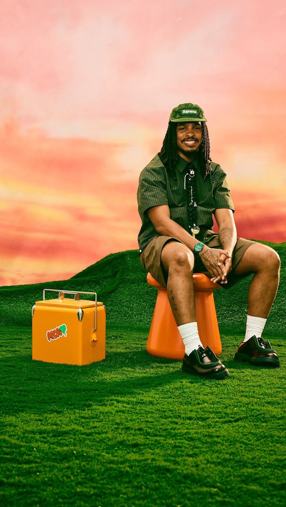 A man with dreadlocks wearing a green cap, dark green shirt, and shorts sitting on an orange stool on green grass with a pastel-colored sky background, next to an orange cooler with a sticker that says "Kega."