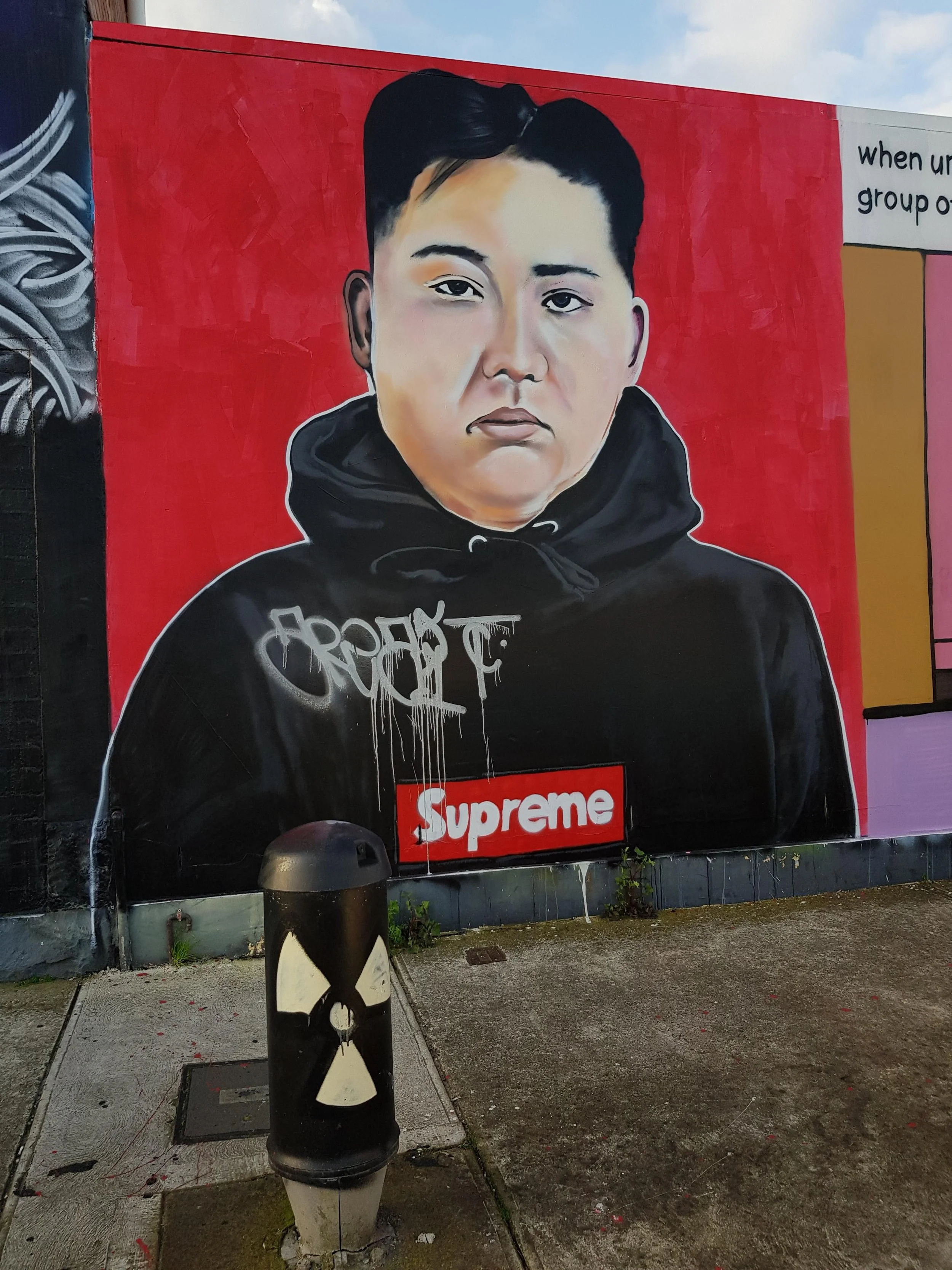 Street art of a person in a black hoodie with 'Supreme' on it, painted on a red wall. A nearby bin has a nuclear symbol.