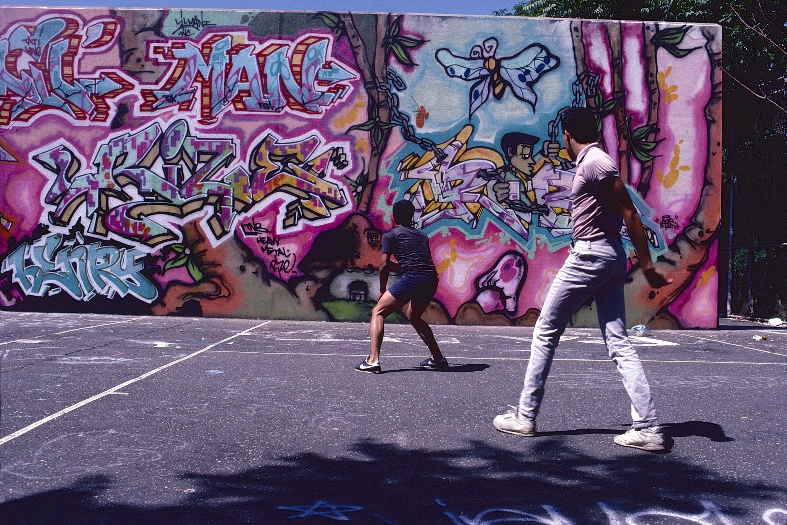 Two people playing on an outdoor sports court in front of a colorful graffiti-covered wall, featuring vibrant designs and lettering.