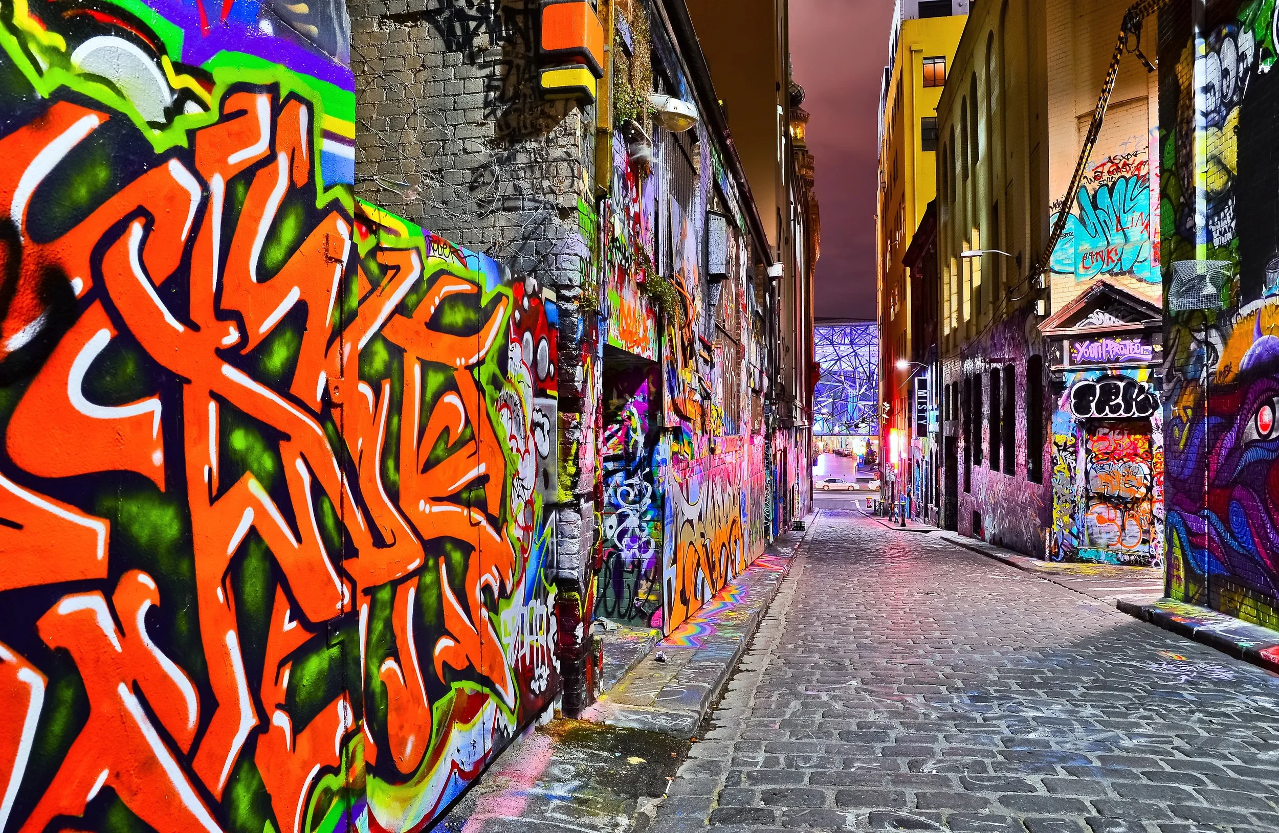 Colorful graffiti-covered alleyway at night with illuminated buildings and a cobblestone path.