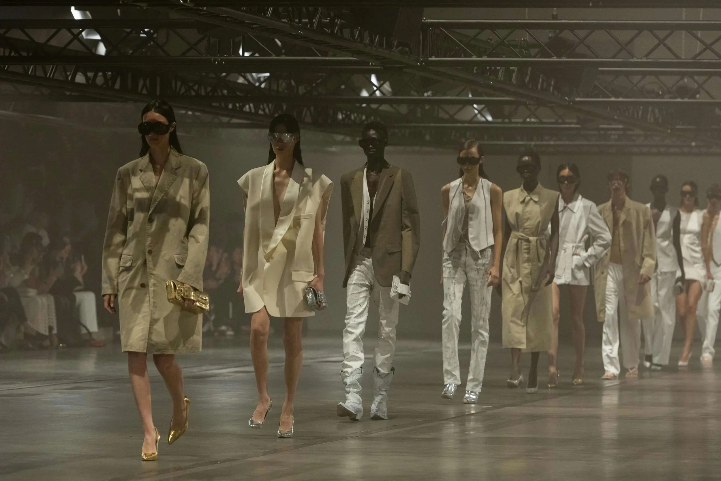 Fashion models walking on a runway during a fashion show, wearing stylish and contemporary outfits in a dimly lit setting.
