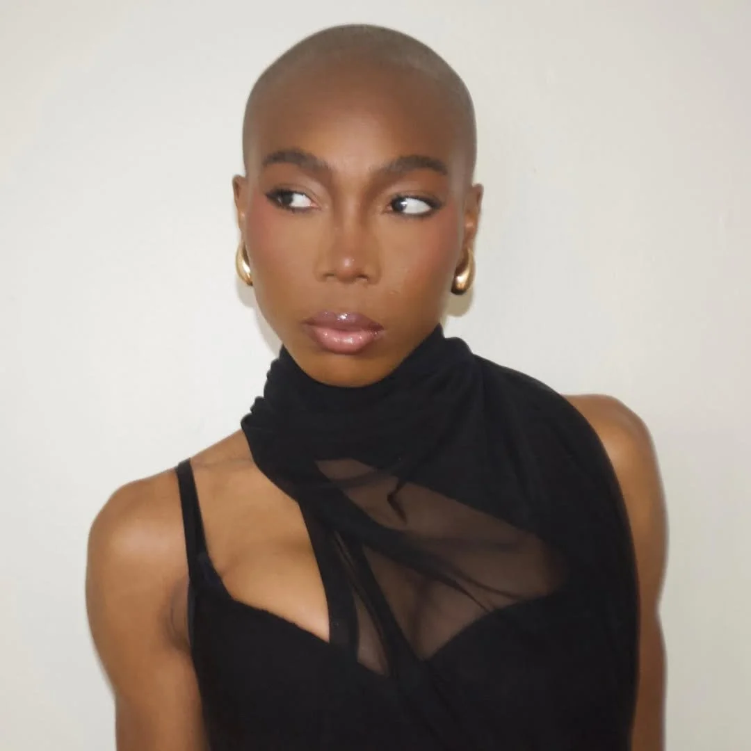 A woman with a shaved head wearing a black top with sheer and cutout details, accessorized with gold hoop earrings.