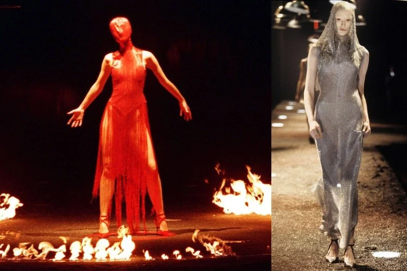 Two models walking on a runway during a fashion show, one in a long, red dress standing near small flames and the other in a metallic silver gown walking on a separate runway.
