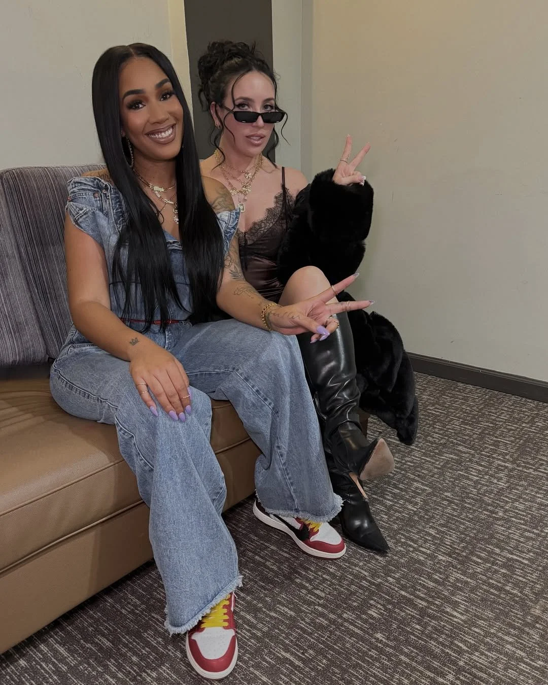 Two women sitting on a couch, both making peace signs. One wears a denim jumpsuit and sneakers, the other in a slip dress and knee-high boots.