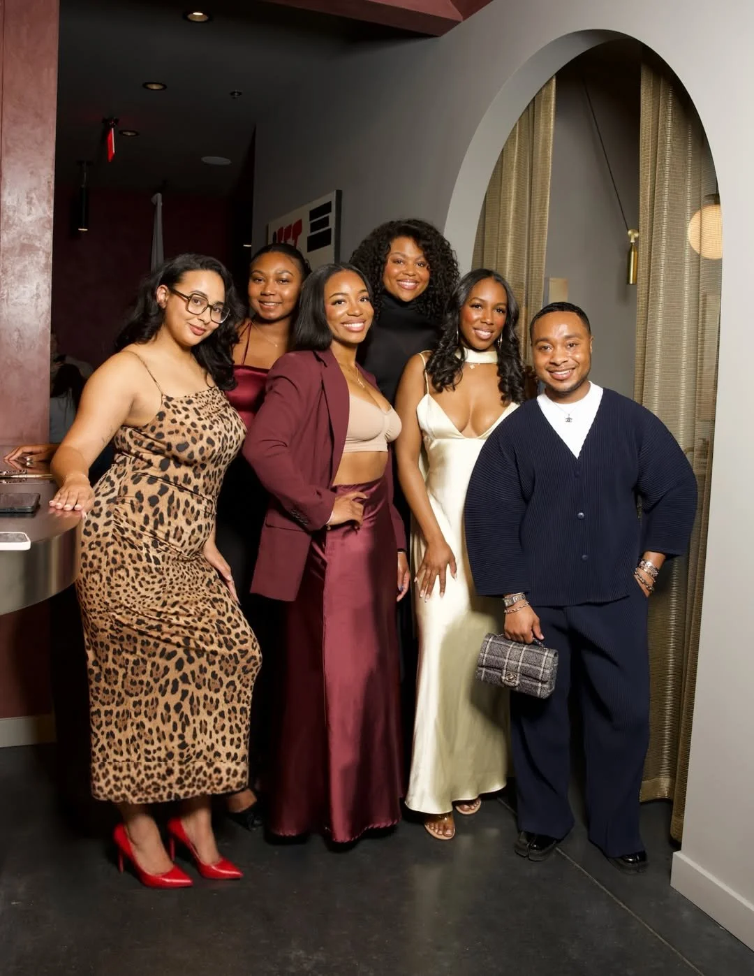 A group of six diverse women and one man dressed in stylish evening attire, posing together indoors at a social gathering or event.
