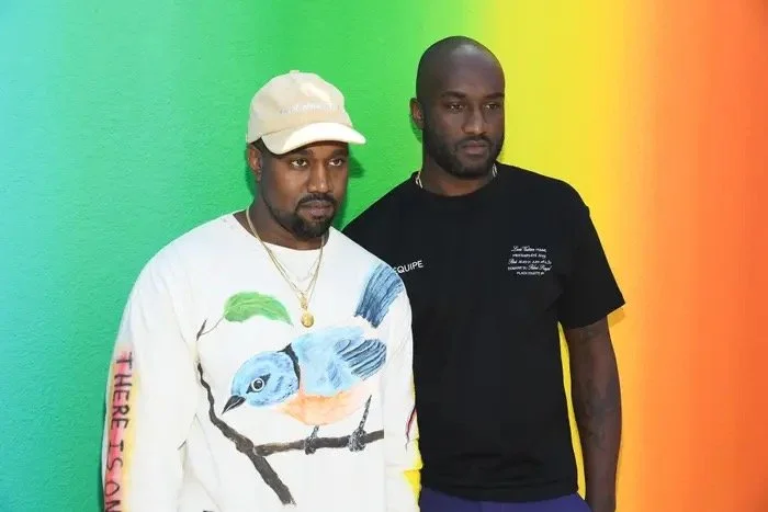 Two men standing in front of a colorful gradient background, one wearing a white sweatshirt with a bird design and a beige cap, and the other wearing a black T-shirt.