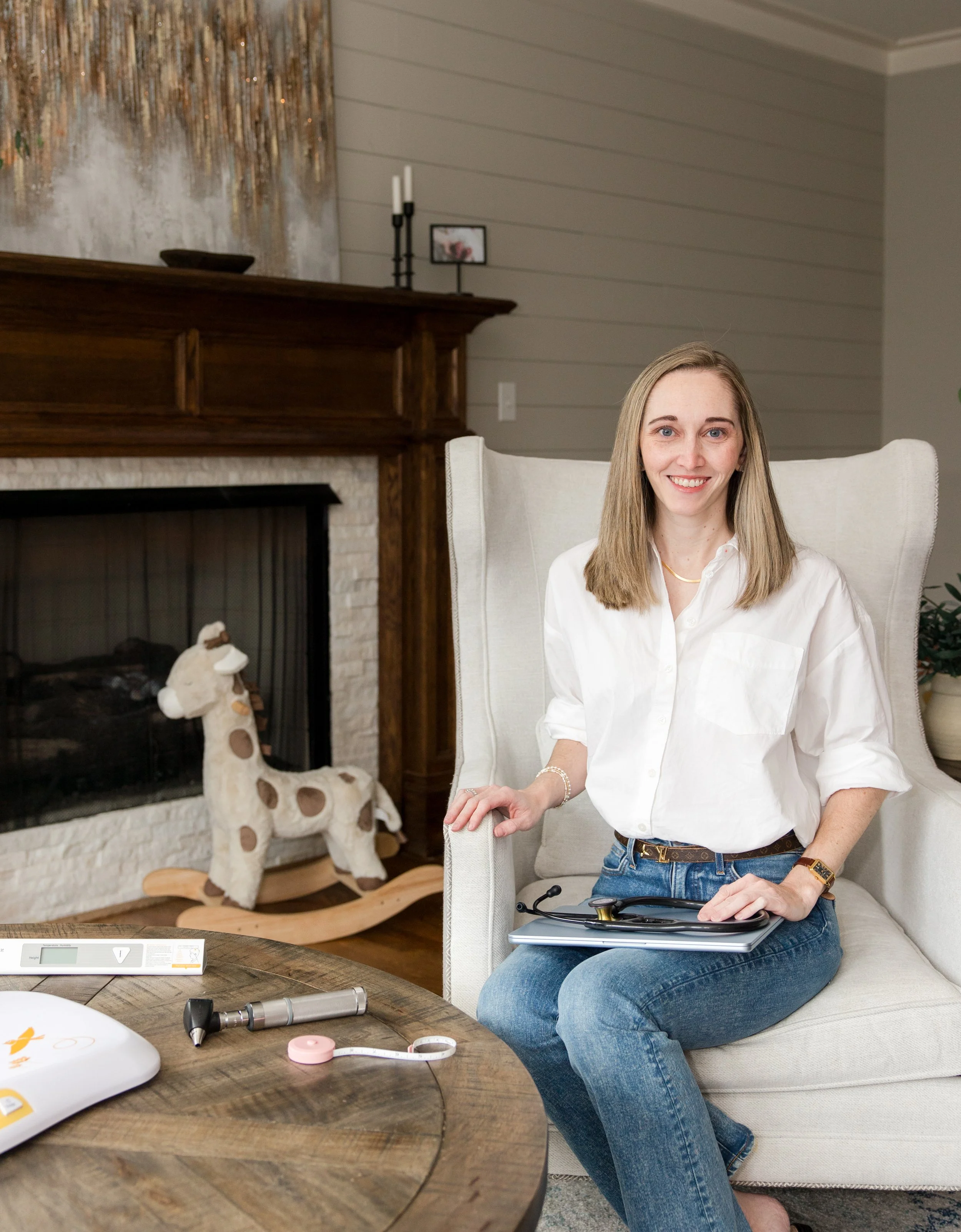 Dr. Laura Mitchell seated in a light armchair holding a stethoscope and tablet, with pediatric tools on a wooden table beside her in a warm home setting.