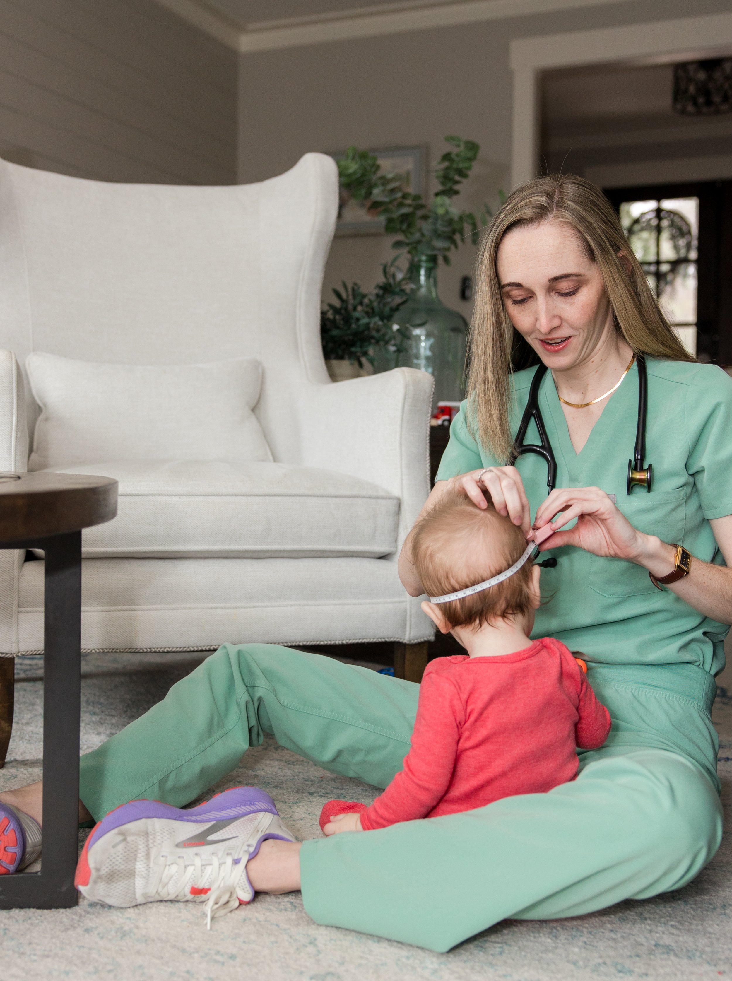 A healthcare professional, wearing green scrubs and a stethoscope, measuring a young child's head with a tape measure indoors.