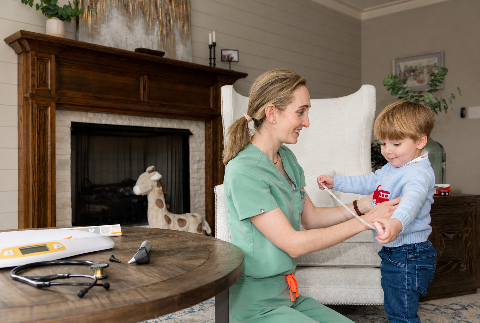 A healthcare professional in scrubs measuring a young boy's chest in a living room. Medical equipment is on the wooden table nearby, and a fireplace with a stuffed giraffe toy in front of it is visible in the background.