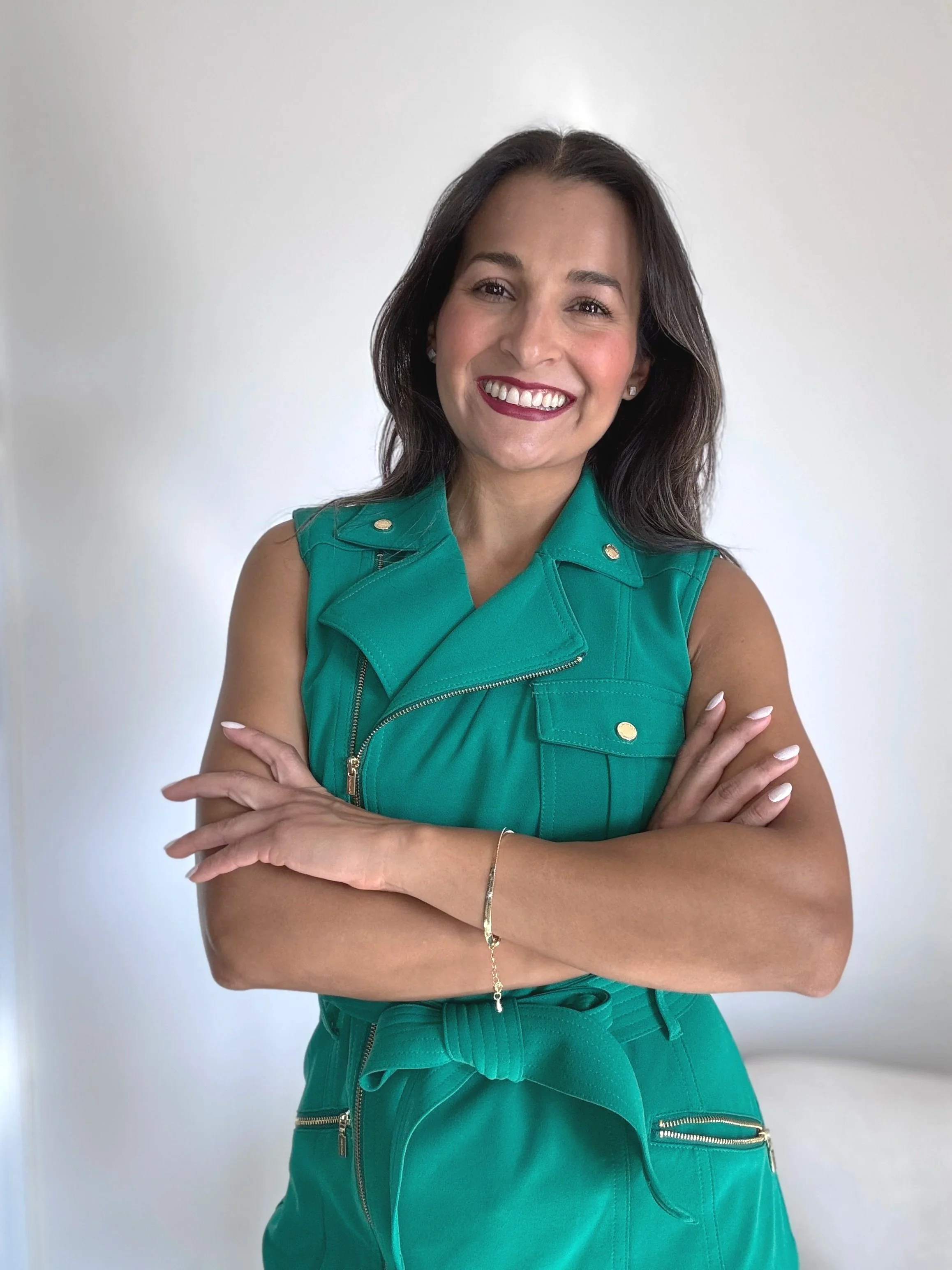 A Residential interior designer woman with dark brown hair, smiling, wearing a sleeveless teal vest with gold buttons and matching teal pants with gold zippers and a tied bow.