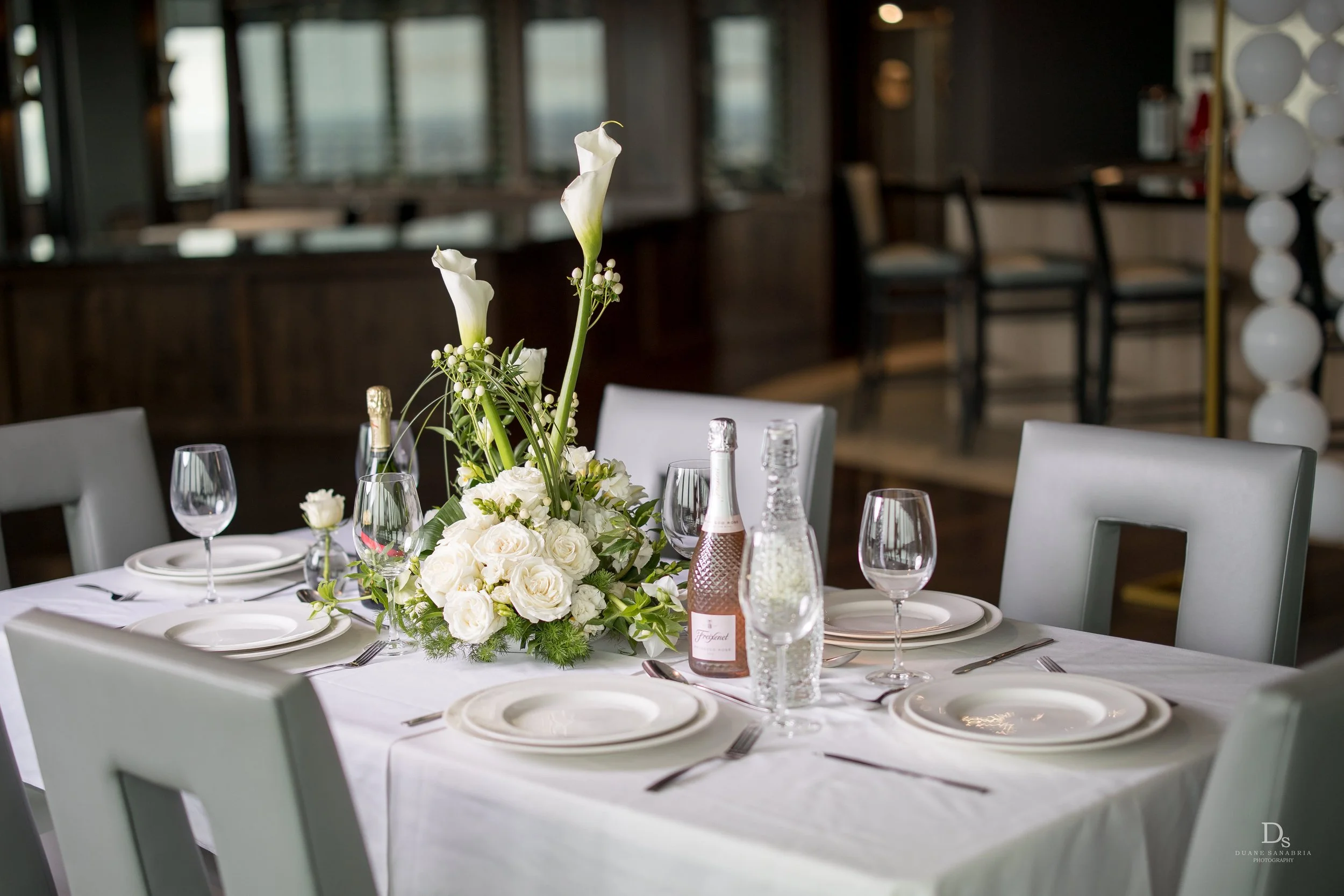 Elegant dining table set with white plates, glassware, silverware, and a floral centerpiece of white roses and calla lilies in a vase, the Penthouse Lounge Slate at Founders Tower