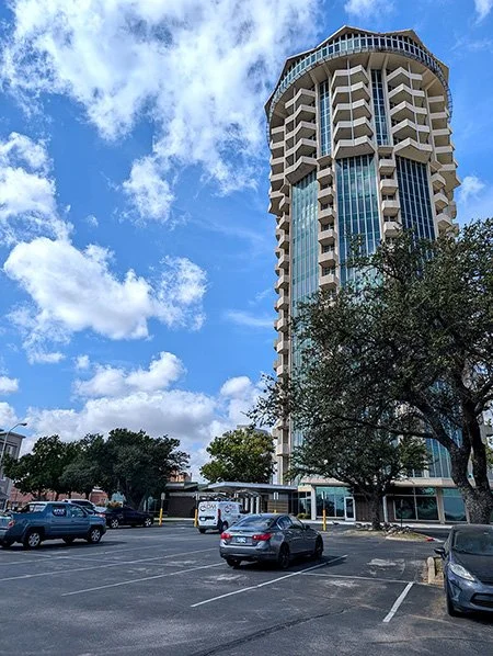 A tall modern building with a circular top section, surrounded by trees in a parking lot with cars and a partly cloudy sky. the Penthouse Lounge Slate at Founders Tower