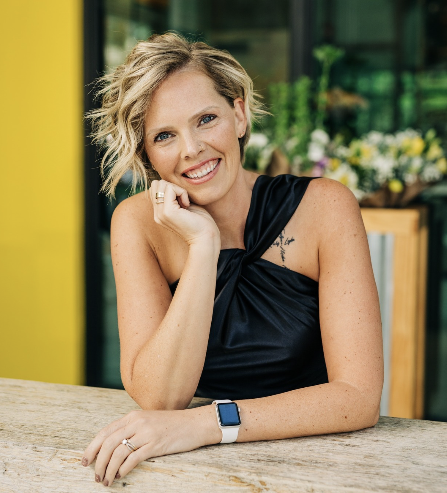 A smiling woman with short, wavy blonde hair, wearing a black sleeveless top, seated at a rustic wooden table with her chin resting on her hand. She has a smartwatch on her left wrist and rings on her fingers. In the background, there are blurred green plants and flowers.