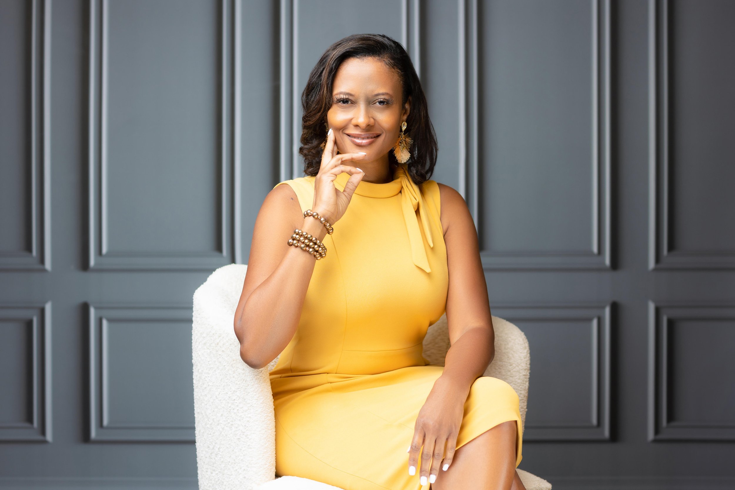 A woman in a yellow sleeveless dress sitting on a white chair in front of a dark gray paneled wall, smiling with one hand touching her face.