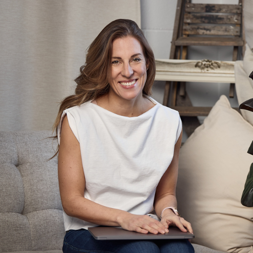 A smiling woman sitting on a sofa with a laptop on her lap, in a cozy, modern living room.