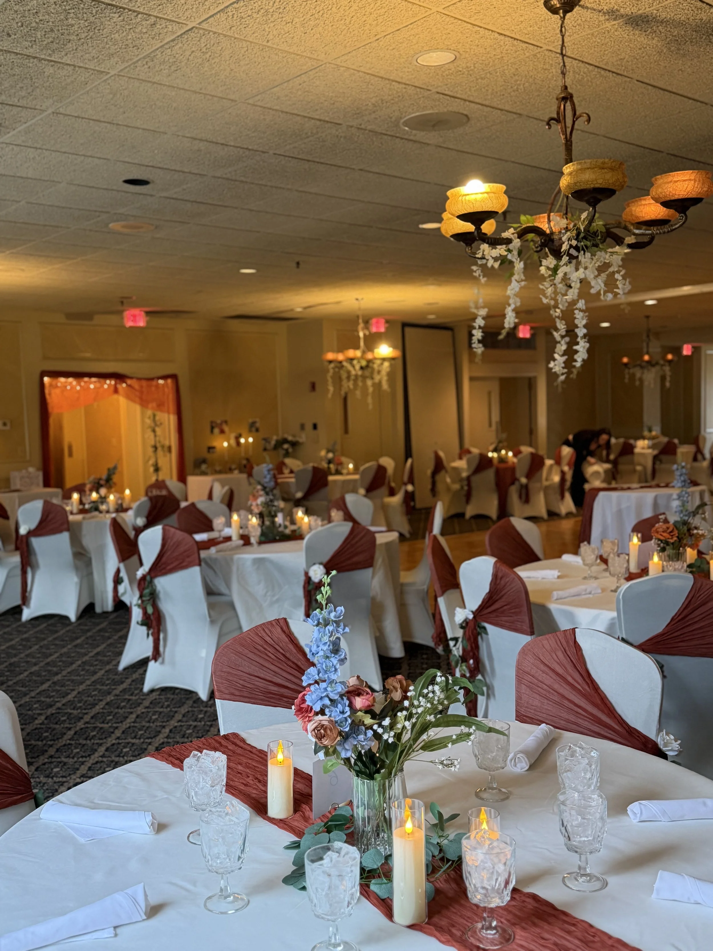 Elegant wedding reception décor with burgundy chair sashes, round tables, candle centerpieces, and hanging floral installations in a ballroom venue