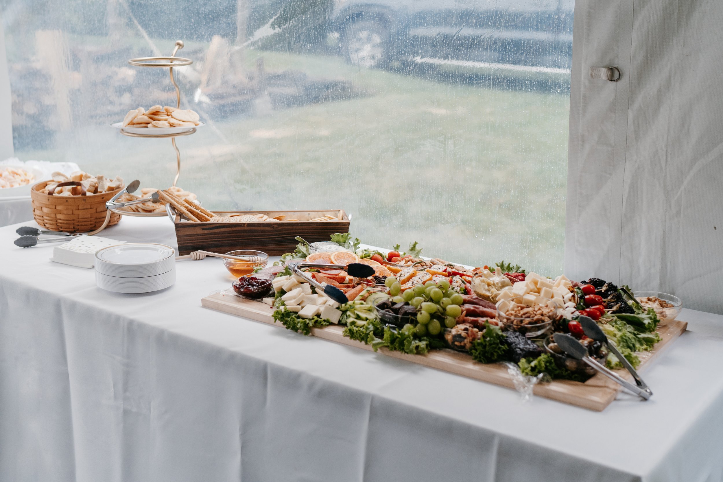 A cheese and charcuterie board with various cheeses, cured meats, grapes, berries, crackers, and garnishes on a wooden slab. A person’s hand is reaching for some cheese.