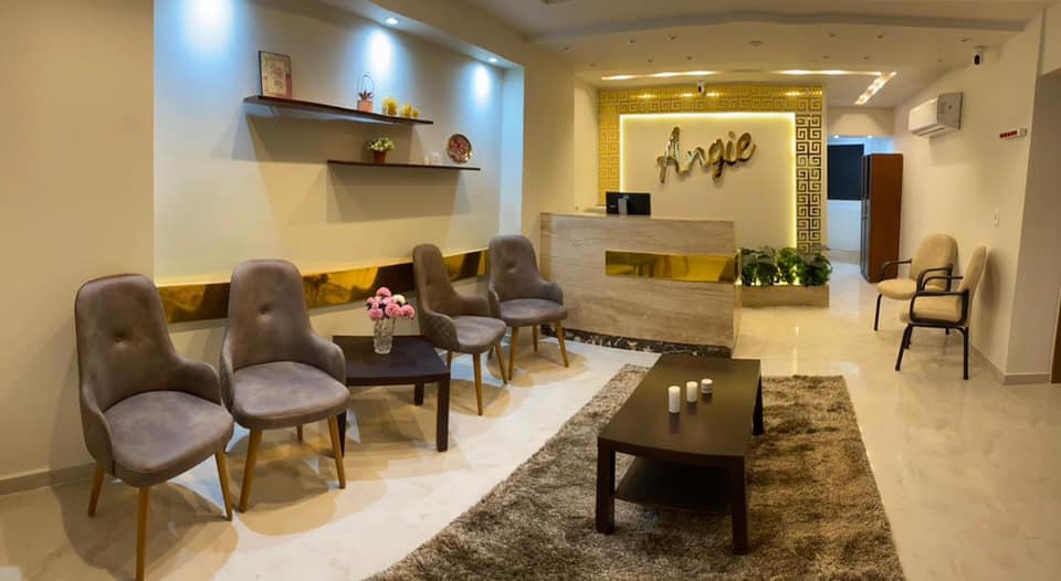 Modern hotel lobby with four gray upholstered chairs, a small table with pink flowers, a brown coffee table with three candles, and a reception desk with a gold and white sign that reads "Angie" behind it. The area has a beige and gold color scheme with decorative shelves on the wall and a seating area to the right.