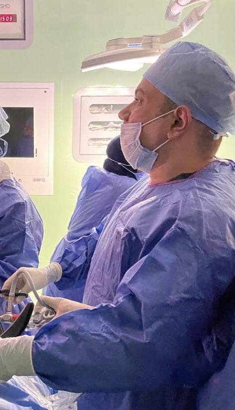 A medical professional in scrubs, a surgical cap, and a face mask, standing in an operating room, with other medical staff and equipment visible in the background.