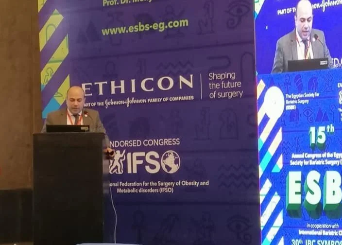 A man in a suit and tie is standing at a podium giving a presentation at a conference. Behind him are large banners with logos and text about ESBS, IFOS, and Johnson & Johnson.