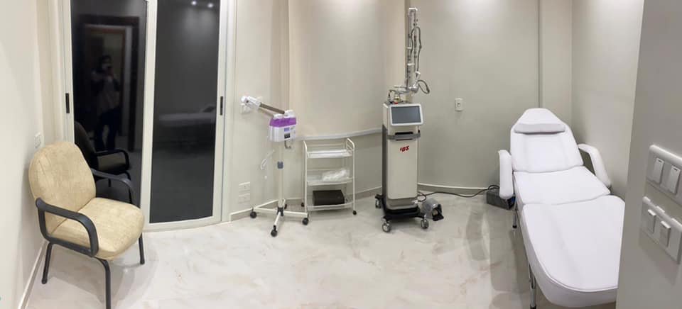 A clean, modern medical examination room with a white examination bed, a beige chair with black armrests, a small medical cart, a medical device on wheels, and a glass door.