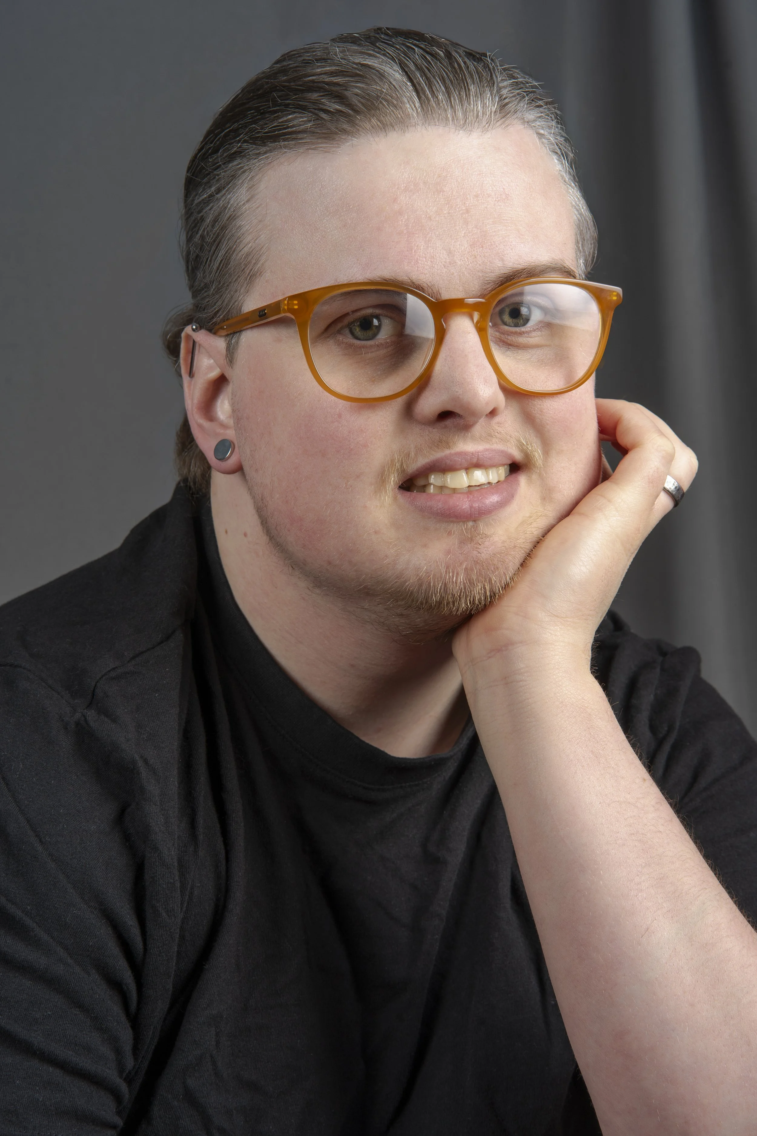 Portrait of a young man with glasses, earrings, and a ring, resting his head on his hand, wearing a black shirt, against a dark gray background.
