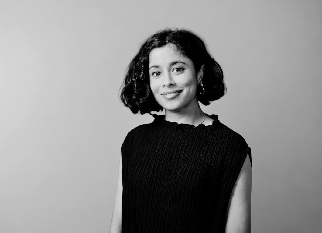 Black and white portrait of a young woman with shoulder-length curly hair, wearing a black sleeveless top, smiling, against a plain background.