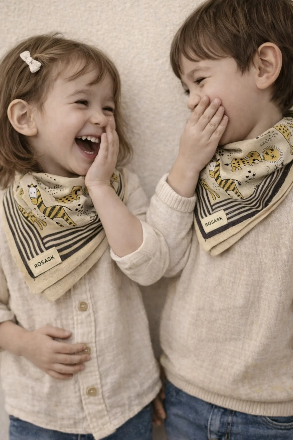 Deux enfants, un garçon et une fille, riant ensemble avec la main sur leur visage, portant des bandanas jaunes avec des dessins d'animaux et des t-shirts beige, contre un mur neutre.
