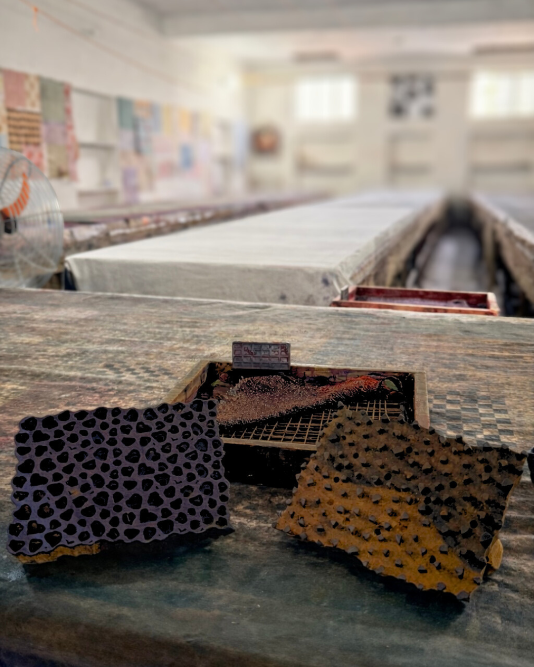 Deux pièces en céramique ou en bois avec des motifs en forme de cœurs et de petites cavités sont posées sur une table en bois. En arrière-plan, on voit une salle lumineuse avec des tables longues recouvertes de tissu ou de papier.