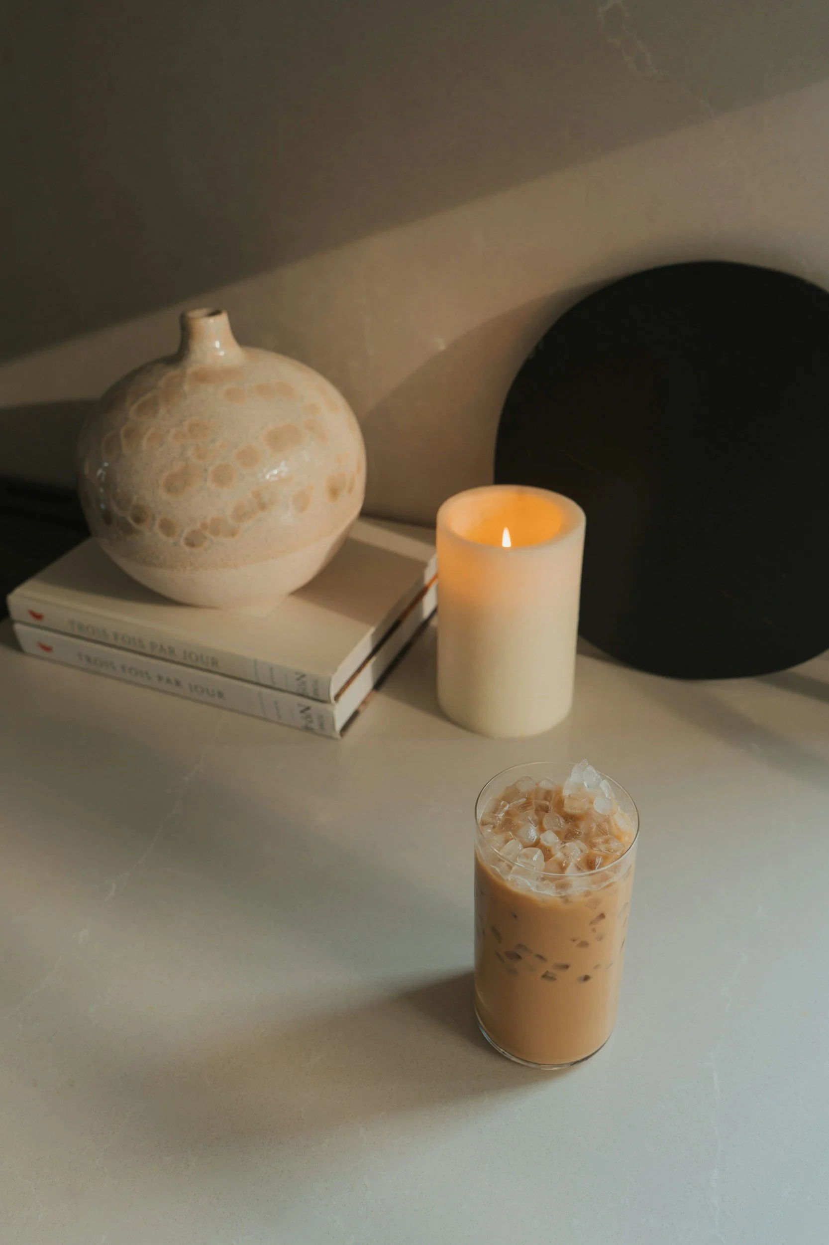 An iced coffee sits on a table with a lit candle and beige objects in the background